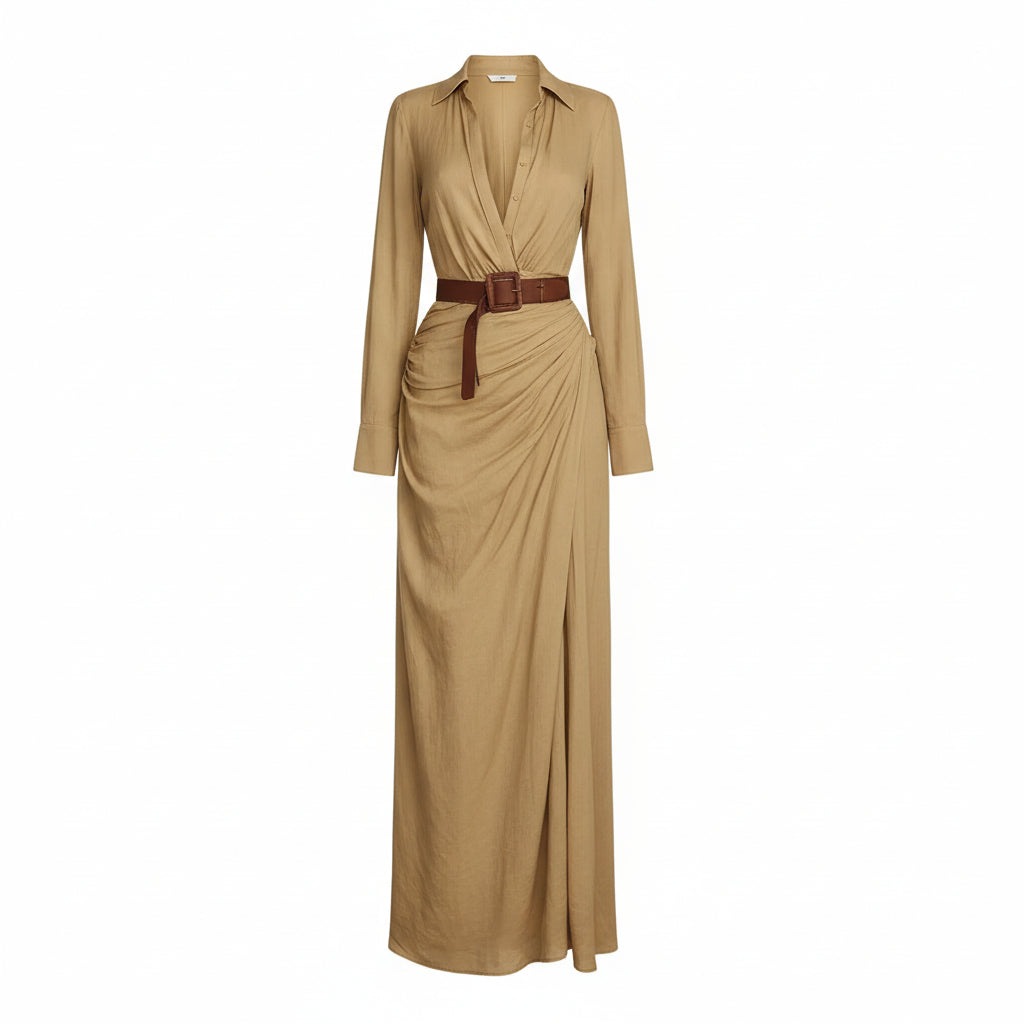 A woman models a flowing tan maxi shirt dress with a deep V-neck, long sleeves rolled to the forearm, and a brown leather belt. She is carrying a woven straw tote bag and wearing flat brown sandals, set against a textured light gray concrete wall.