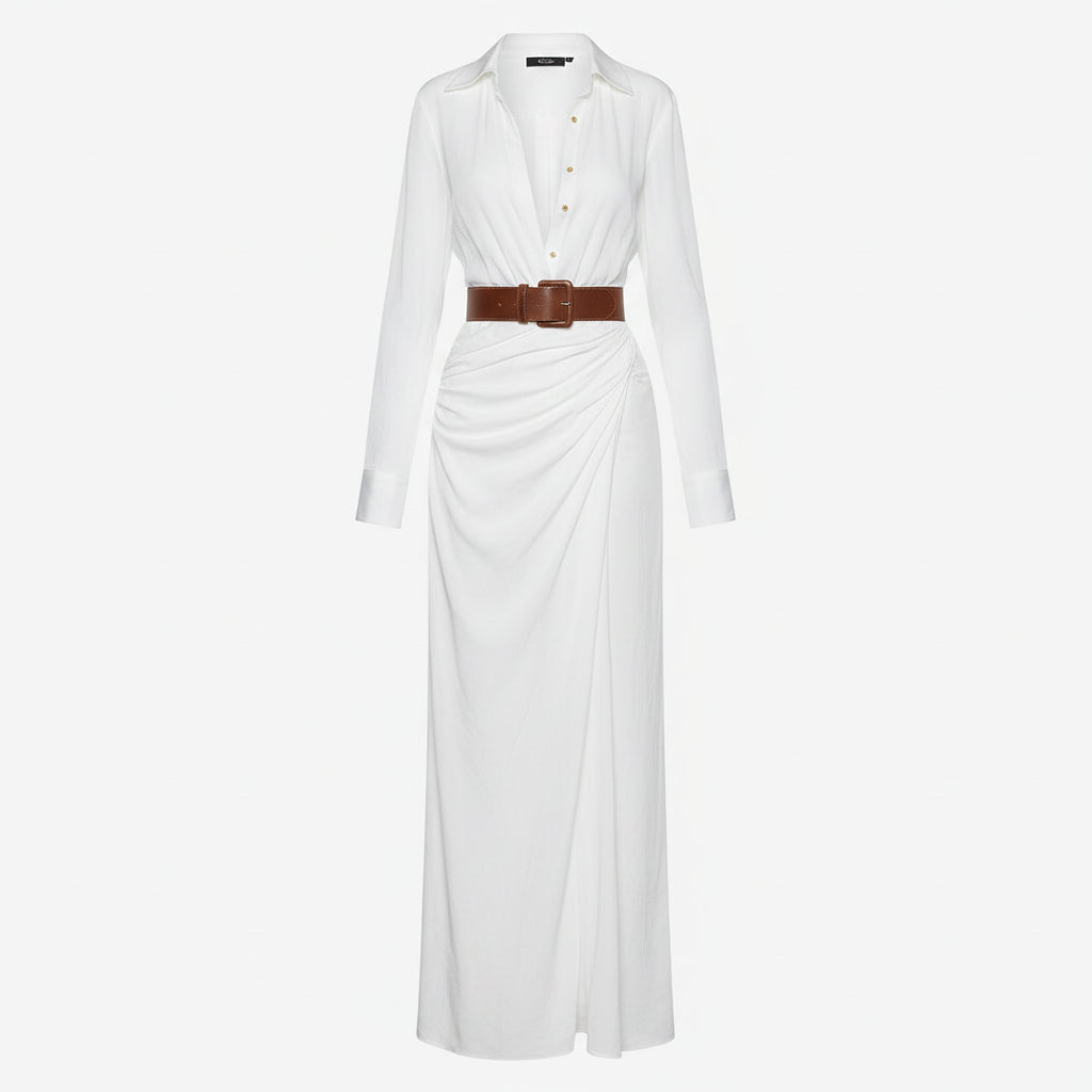 A woman models a white long-sleeve wrap-style maxi dress with a deep V-neckline, tucked into a matching white skirt, styled with a brown belt and a woven straw tote bag against a textured light gray wall.